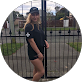 Woman in a black dress and black cap standing outdoors in front of a metal fence and greenery.