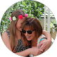 Smiling woman wearing sunglasses hugs a young girl with a pink flower headband in a bright outdoor setting.